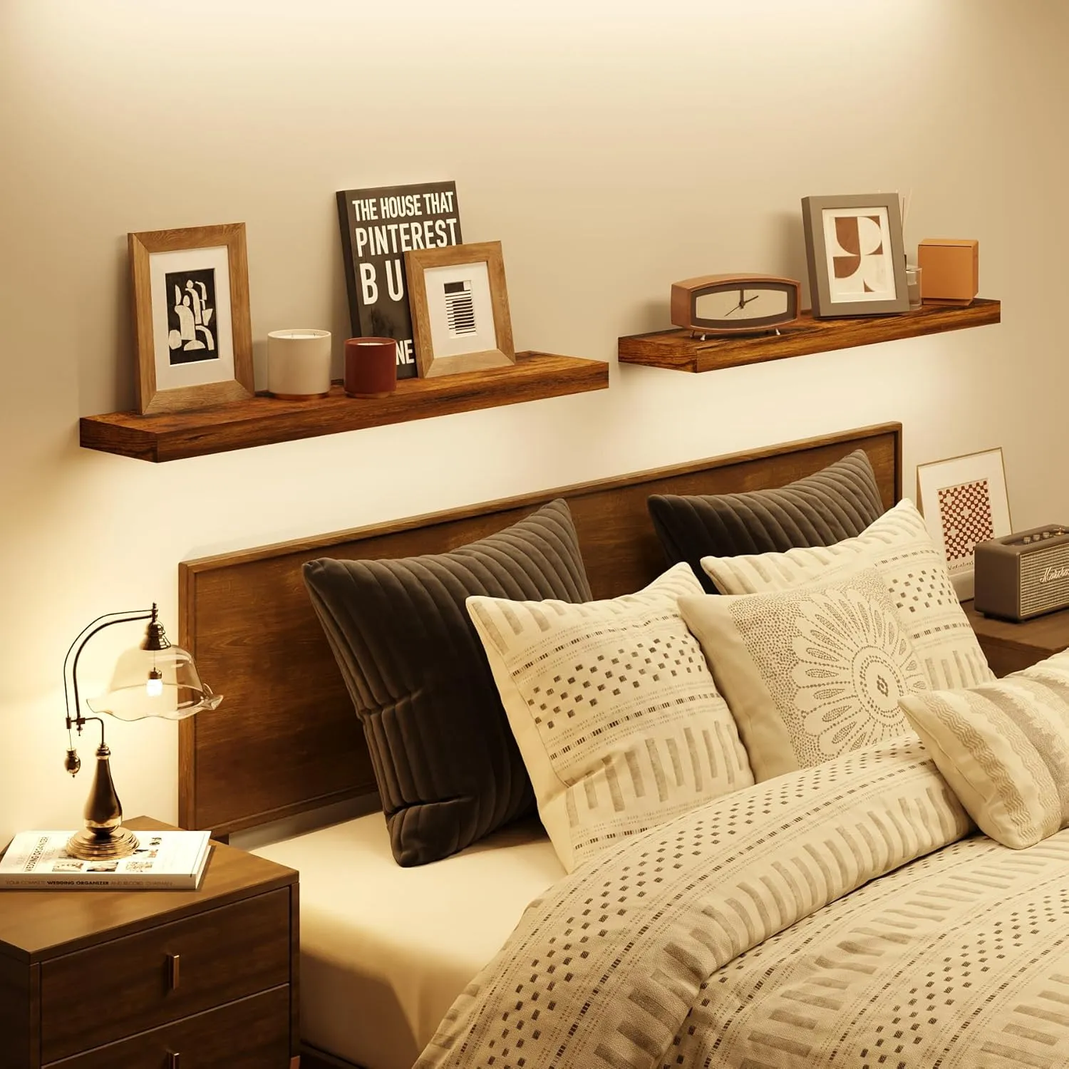 This is a cozy, modern bedroom corner. Above the light-colored wooden headboard, there are two layers of floating shelves installed, displaying delicate items such as photo frames, an alarm clock, and green plants. On the left side, there is a dark wooden bedside table paired with a black table lamp and books. The overall decor features beige bedding paired with gray throw pillows, complemented by warm-toned walls and wooden elements, creating a tranquil and comfortable resting atmosphere.
