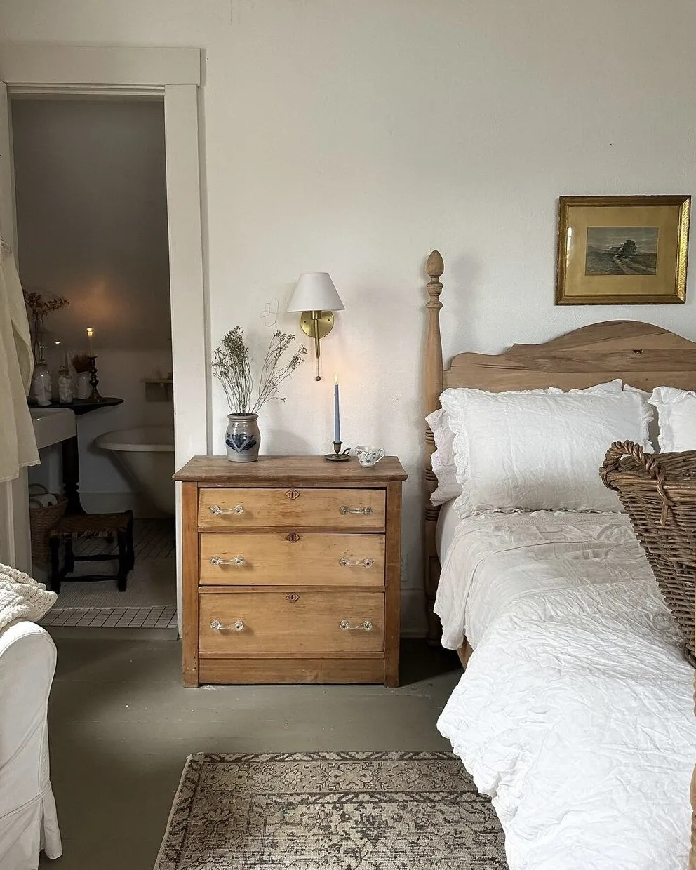 This serene bedroom centers around a wooden bed dressed in crisp white linens, accented by a simple wooden nightstand. A vase with fresh greenery and a candle adds a natural touch, while a gold-framed landscape painting and a minimalist wall lamp create a soft, ambient glow. The open view to the adjoining bathroom with a freestanding tub enhances the airy, relaxed feel. Warm wood tones and neutral colors establish a calming and inviting retreat.