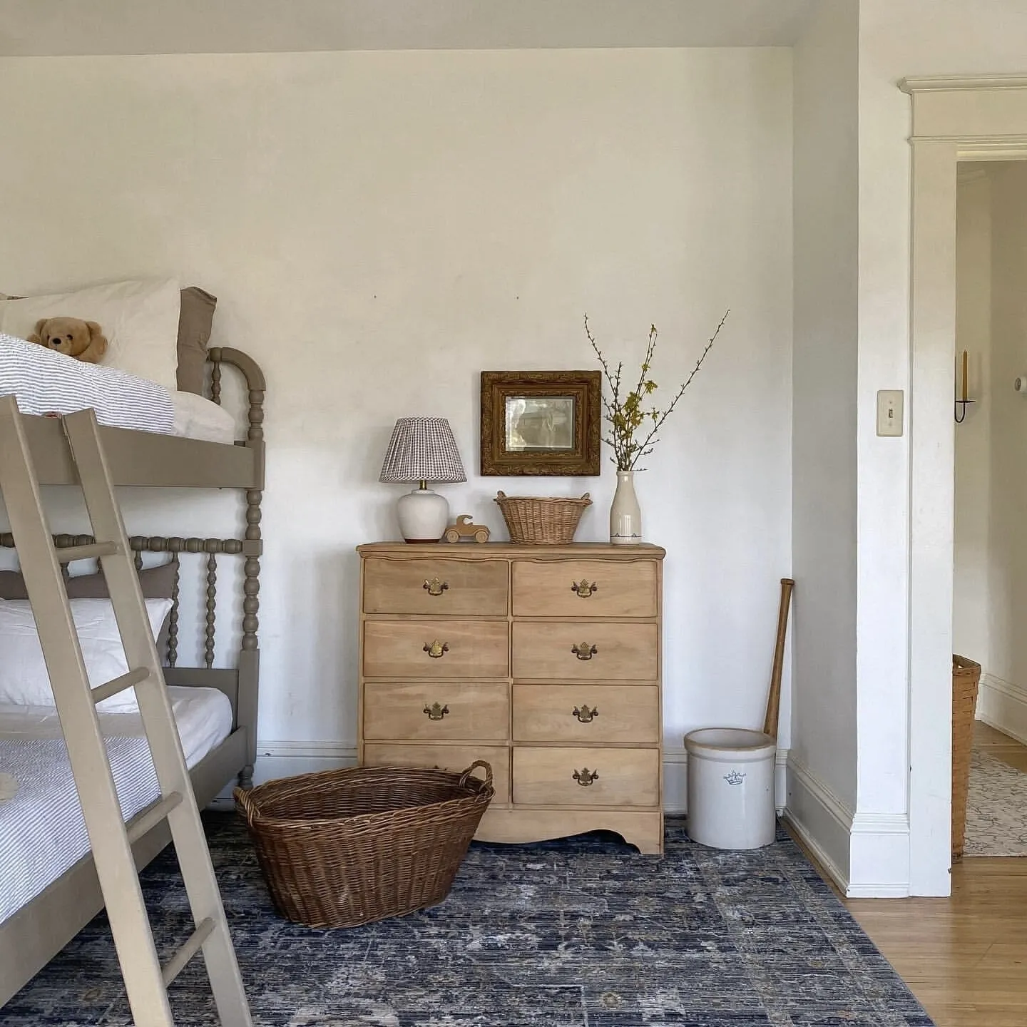This cozy bedroom features a soft grey bunk bed with a plush teddy bear on the upper level. A light wood dresser holds a lamp, small trinkets, a photo frame, and a vase with delicate branches. A blue-grey patterned rug anchors the space, complemented by a woven basket and simple white storage. The white walls and natural light create a calm, orderly, and soothing retreat.