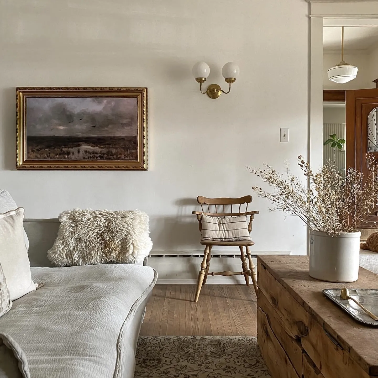 This cozy living room features a soft gray sofa with plush pillows, paired with a wooden armchair holding a striped cushion. A landscape painting in a gold frame and a matching sconce adorn the white wall. A wooden side table displays a large vase of dried flowers on a tray, while a patterned rug ties the space together. The warm, neutral palette creates a harmonious and inviting atmosphere.