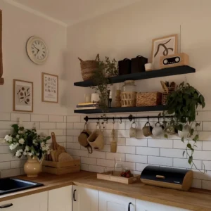 A cozy kitchen corner with light-toned walls and wooden countertops. Two black shelves display woven baskets, potted greens, decorative jars, and a small radio, adding rustic charm. A circular clock and minimalist art hang above, while mugs and a vase with white flowers sit below. The white tile backsplash keeps the space fresh and clean, creating a warm, inviting feel.